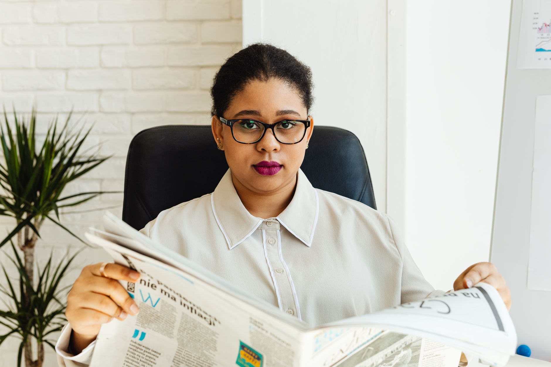 Professional woman with glasses reading a newspaper at her office desk, surrounded by modern decor. — Frequently Asked Questi
