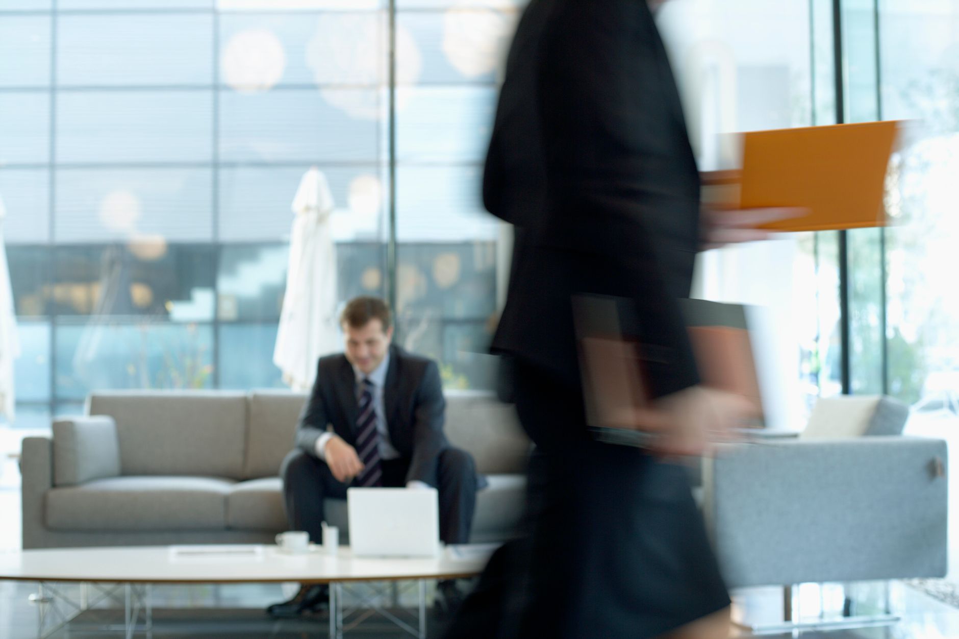 Businessman sitting in modern office lounge using a laptop while colleague walks past, creating dynamic blur. — How To Get A
