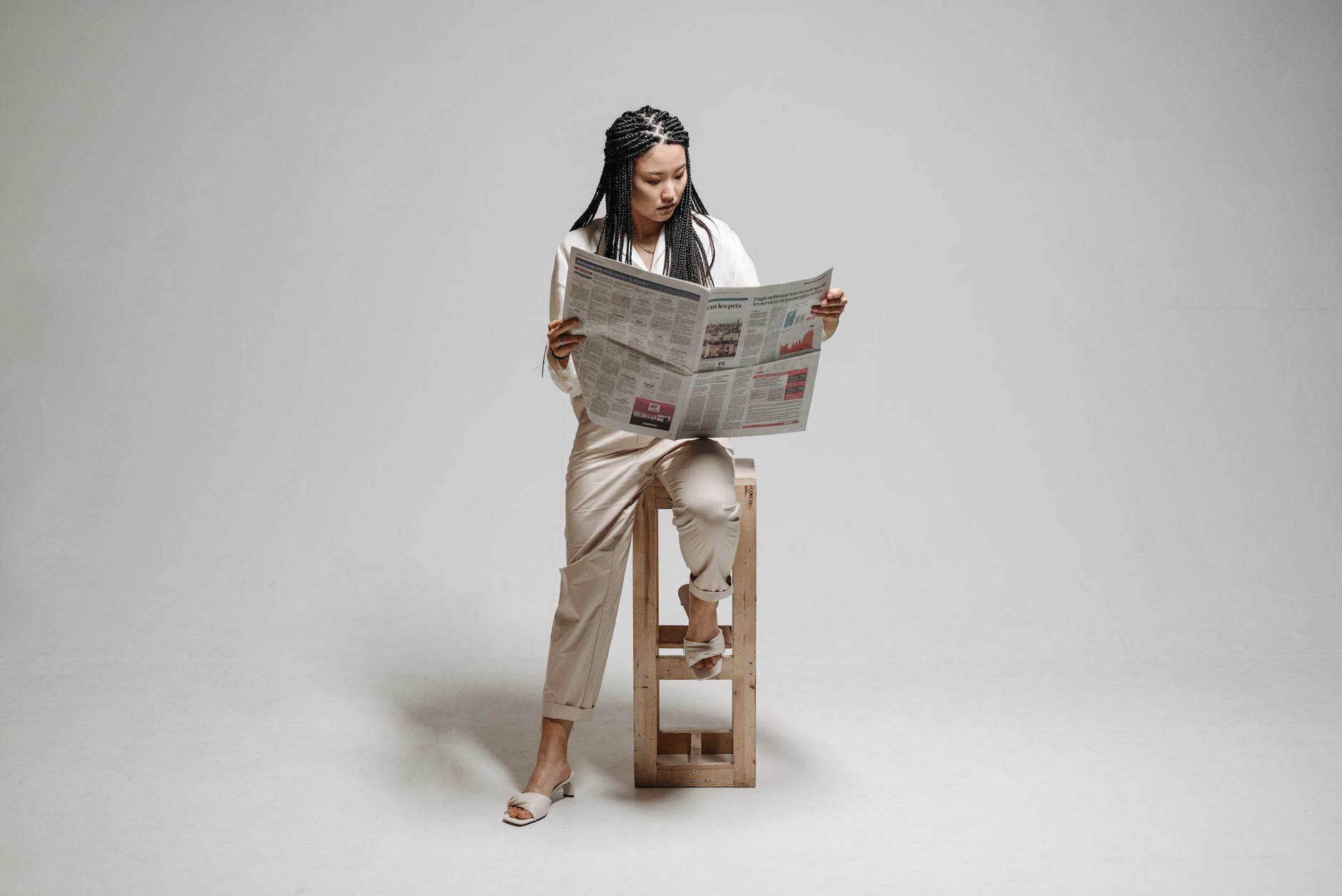 A woman sits on a stool with a braided hairstyle, reading a newspaper in a minimalistic studio. — Sports Streams: The Factors