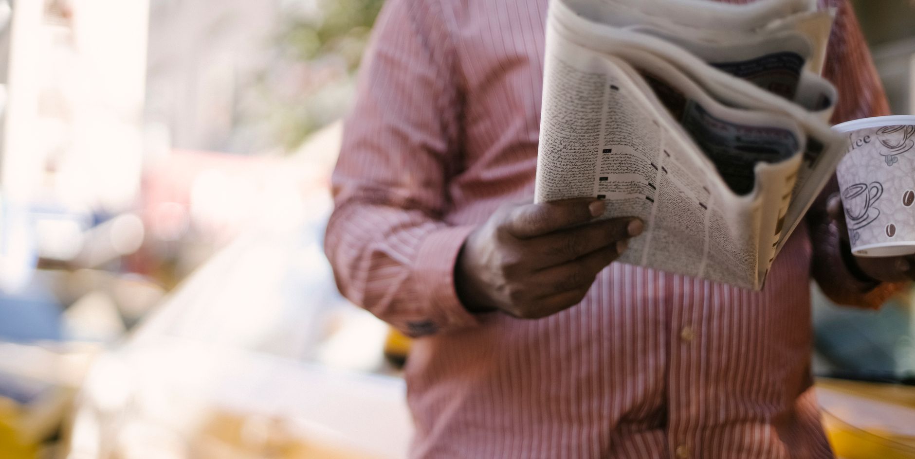 Crop unrecognizable male reading newspaper and holding paper cup with hot beverage while standing on city street — Ba