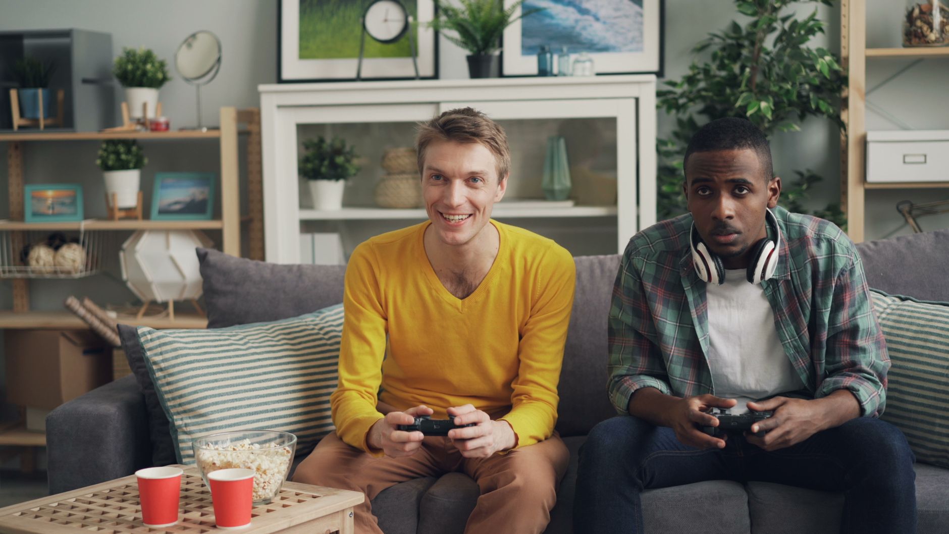 Two friends playing video games together on a sofa, one holding a controller and the other wearing headphones. — W88 Sports B