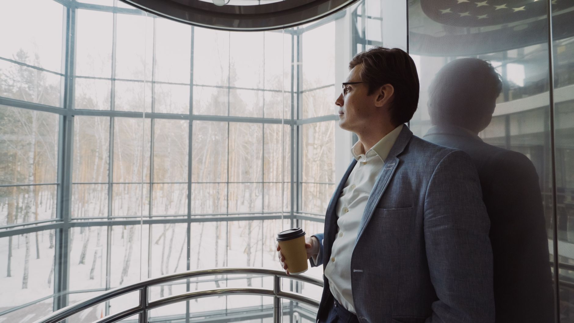 Businessman in blue suit with coffee cup looking out a large window, reflecting on a winter landscape. — What You Should Know