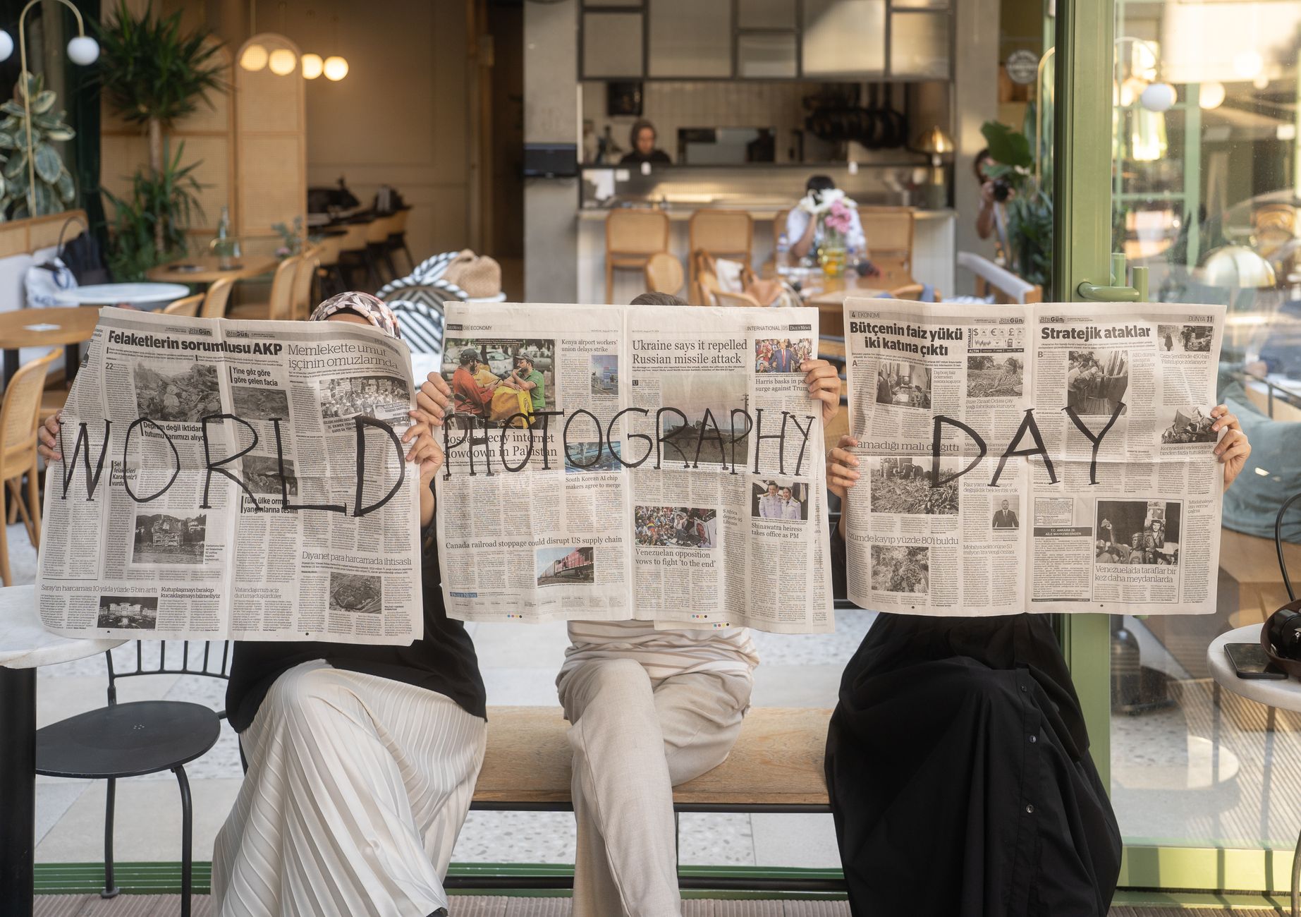 Three people holding newspapers promoting World Photography Day in a cozy Ankara café. — Why You Should Use the Cosmostation